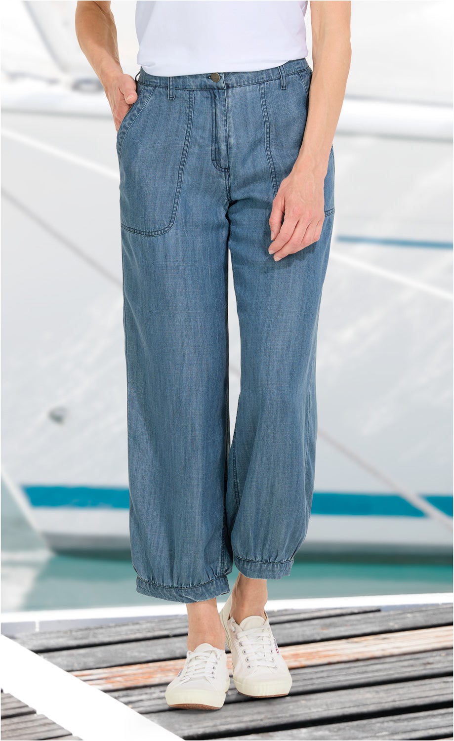 Person trägt blaue Lyocell-Hose mit weitem Bein und elastischem Saum, weiße Sneaker, Hände in den Taschen, Hintergrund mit unscharfem Wassersportboot und Steg