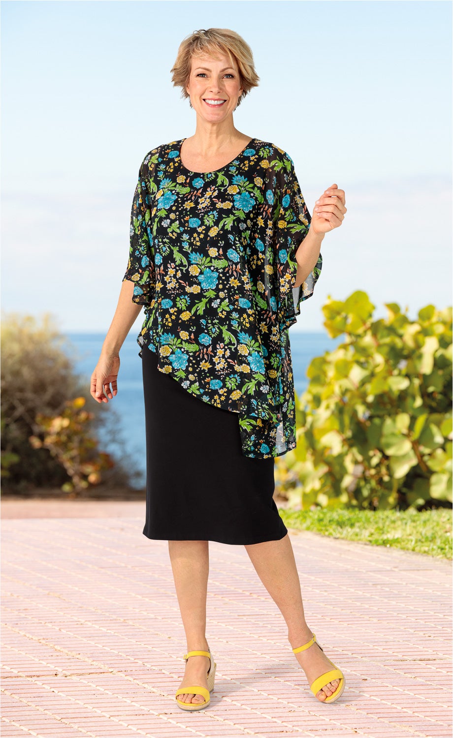 Frau in schwarzem Viskose-Kleid mit buntem Blumenmuster, gelben Sandaletten, steht auf gepflastertem Weg, im Hintergrund Meer und grüne Büsche, bei sonnigem Himmel