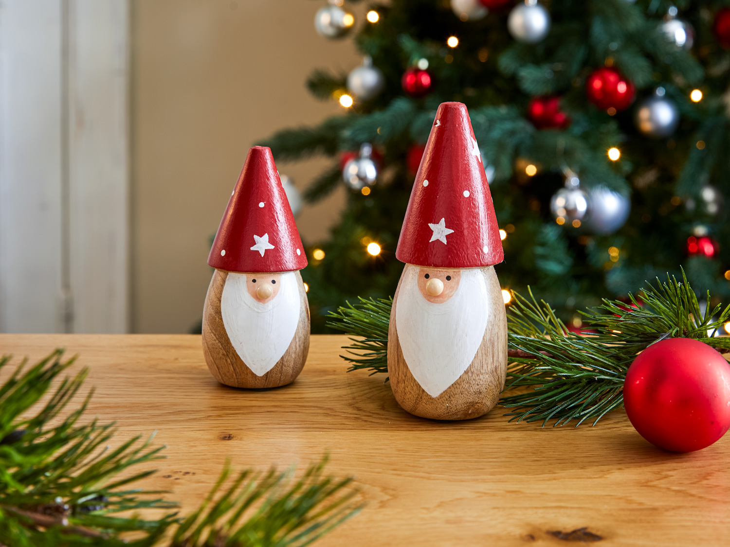 Zwei hölzerne Nikolausfiguren mit roten Zipfelmützen und weißen Bärten auf Holztisch, Weihnachtsbaum mit roten und silbernen Kugeln im Hintergrund, Tannenzweige und rote Weihnachtskugel dekorativ platziert