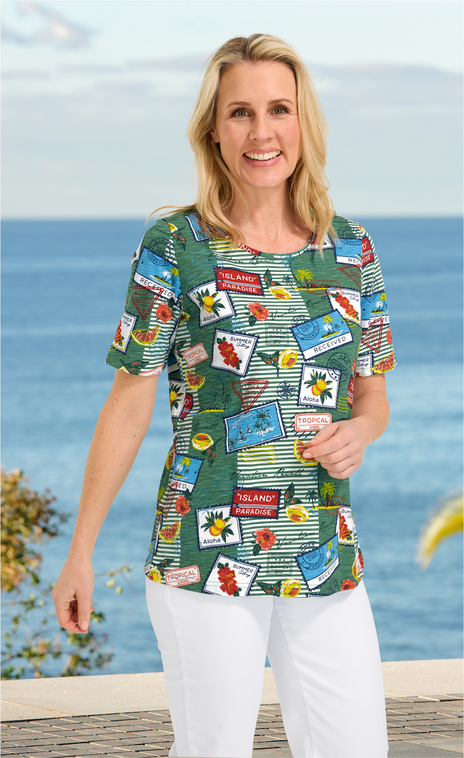 Frau trägt buntes Baumwoll-Shirt mit tropischem Muster und weißen Hosen, Hintergrund Strand und Meer, lächelt in die Kamera