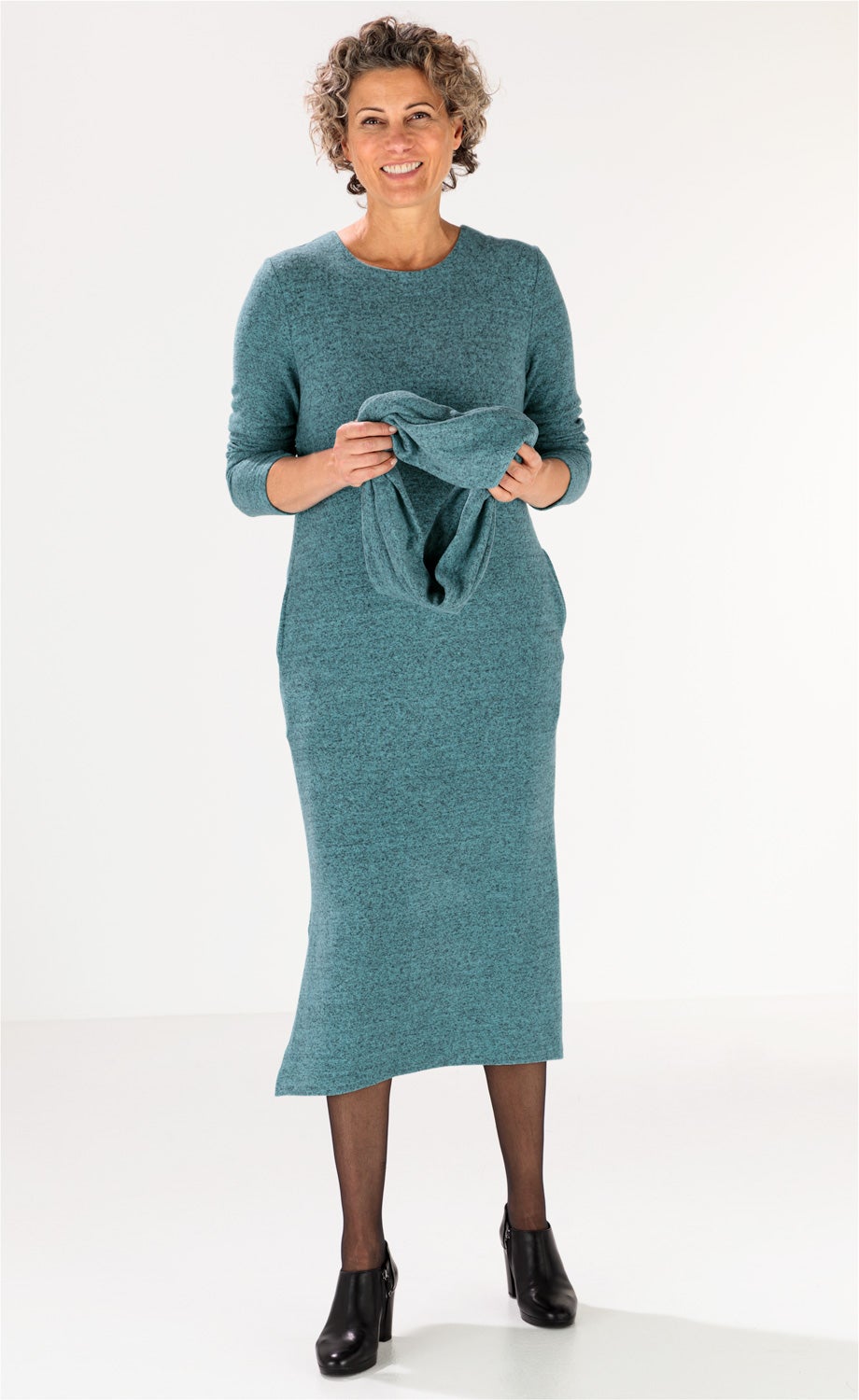 Frau trägt langes, türkisfarbenes Viskose-Mix-Kleid mit langen Ärmeln, hält passenden Schlauchschal, kombiniert mit schwarzen Strumpfhosen und schwarzen, hochhackigen Stiefeletten, freundliches Lächeln, helle Hintergrundfarbe