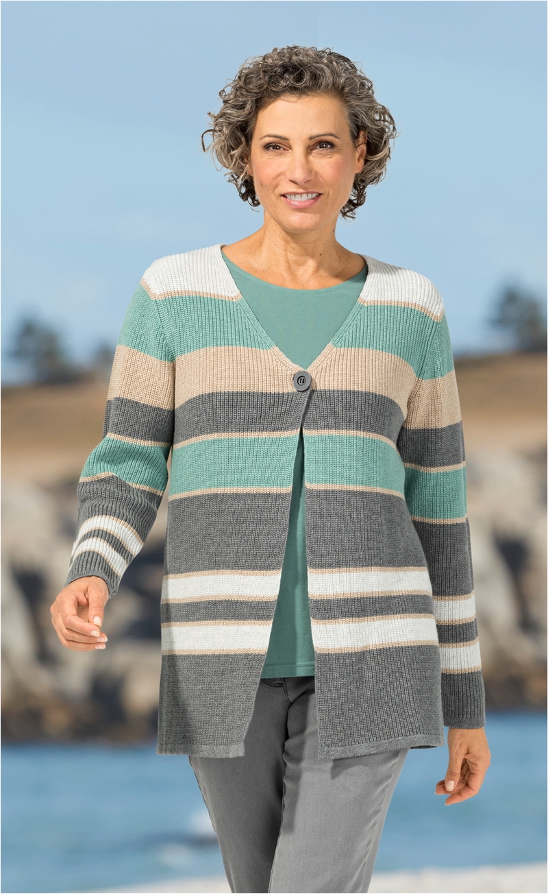 Frau trägt Baumwoll-Strickjacke mit breiten Streifen in Grau, Türkis, Beige und Weiß, einfarbiges Türkis-Shirt, graue Hose, lockige kurze Haare, lächelt, stehend vor Strandkulisse mit Felsen und Meer im Hintergrund