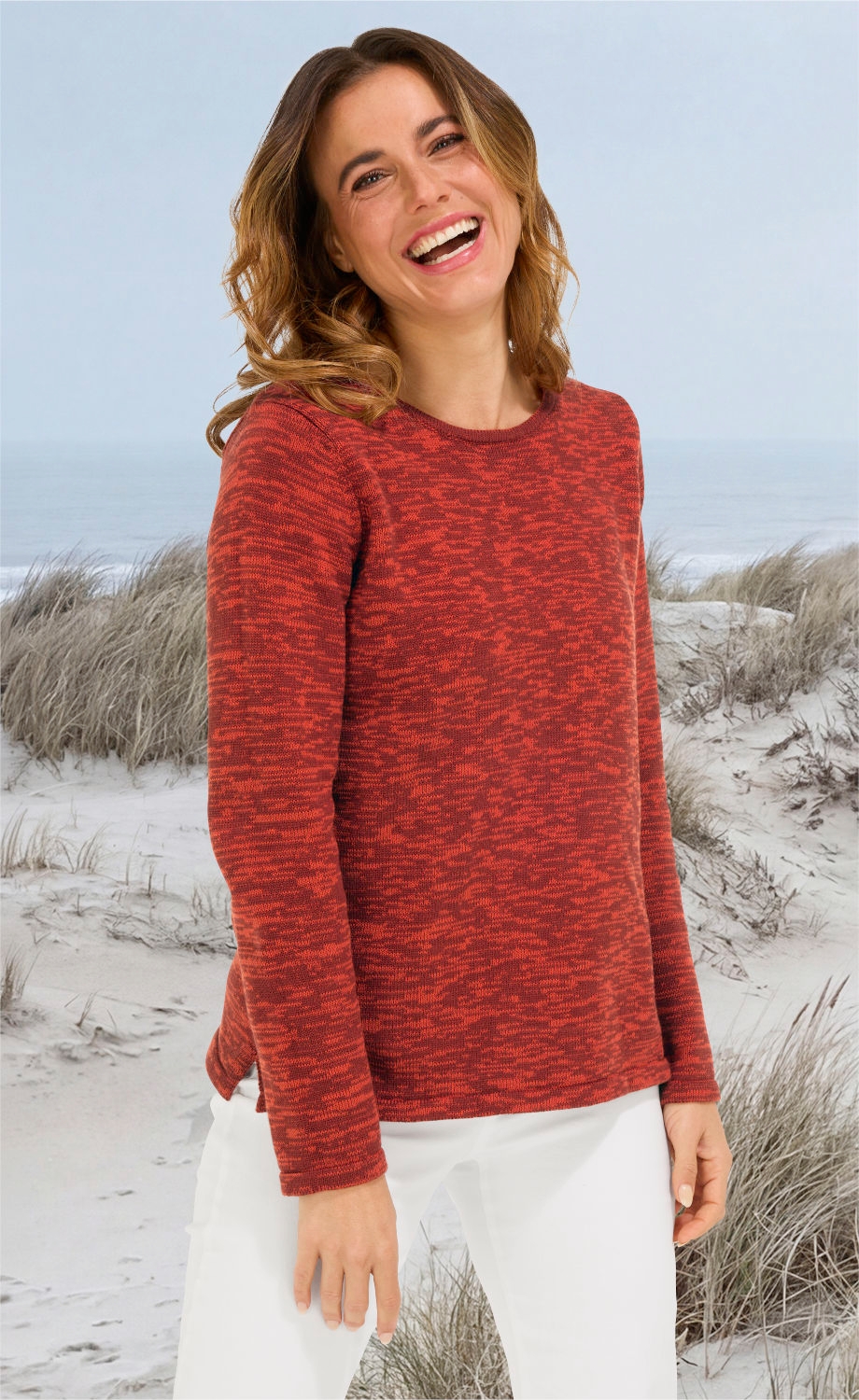 Frau am Strand trägt roten gemusterten Baumwoll-Pullover mit rundem Ausschnitt und weiße Hose, lächelt, halbe Frontansicht, Hintergrund Dünen und Meer bei bewölktem Himmel.