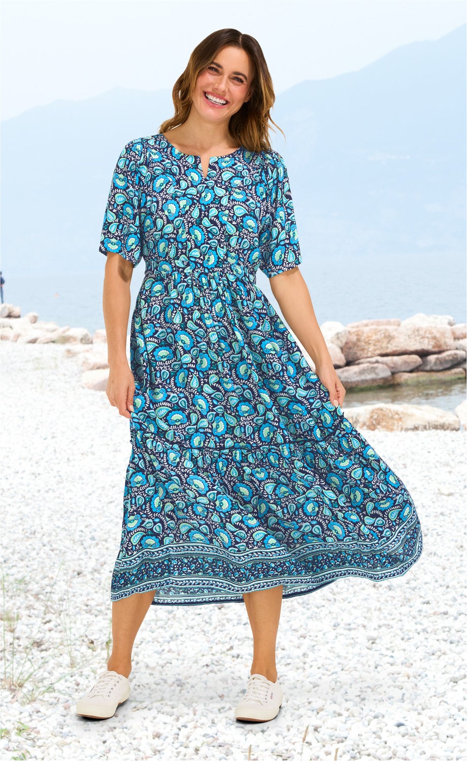 Frau am Kiesstrand trägt blaues Viskose-Kleid mit floralem Muster, halblangen Ärmeln und Knopfleiste, kombiniert mit weißen Sneakern, lächelnd vor Berg- und Seehintergrund