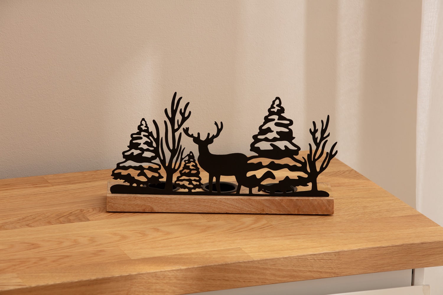 Teelichthalter mit schwarzem Metallmotiv aus Tannenbäumen, kahlen Bäumen und Hirsch, auf rechteckigem Holzsockel, stehend auf hellem Holztisch vor beigem Hintergrund