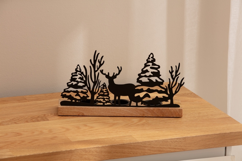 Teelichthalter mit schwarzem Metallmotiv aus Tannenbäumen, kahlen Bäumen und Hirsch, auf rechteckigem Holzsockel, stehend auf hellem Holztisch vor beigem Hintergrund