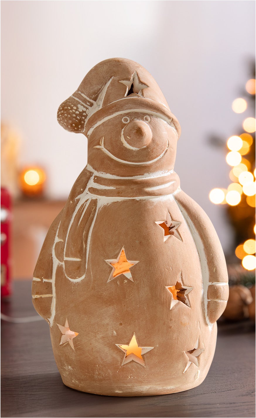 Terracotta Windlicht in Schneemannform mit Sternausschnitten, beleuchtet von innen, trägt Mütze und Schal, auf dunklem Tisch vor unscharfem, warm beleuchtetem Hintergrund mit Weihnachtsdeko.