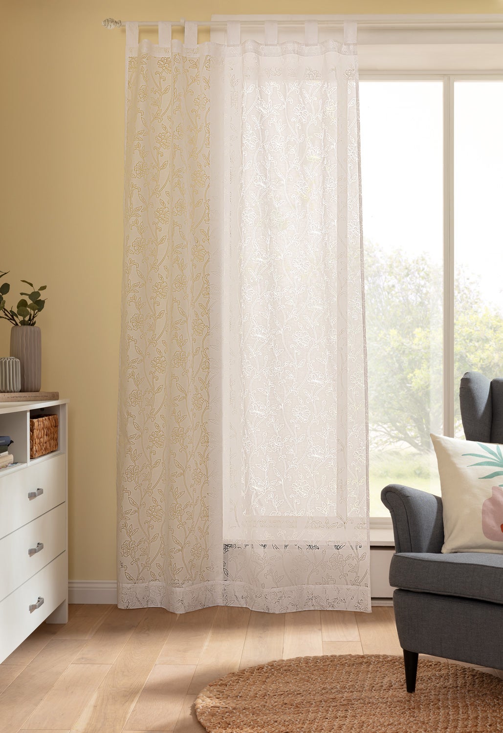 transparenter weißer Vorhang mit floralem Muster auf Schlaufen an Fenster, Tageslicht, helles Wohnzimmer mit grauem Sessel, Kissen mit Blumenmuster, weißem Sideboard, Holzfußboden und rundem Juteteppich