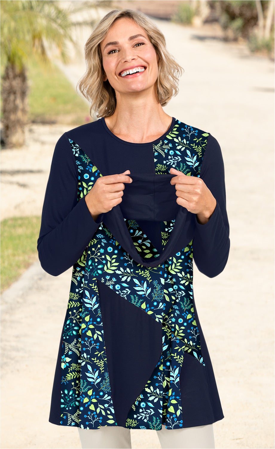 Frau trägt marineblaues Viskose-Mix-Shirt mit floralen Mustern in Blau- und Grüntönen, hält vorne den Kragen mit beiden Händen, lächelt, steht auf hellem Weg mit unscharfem Grün im Hintergrund