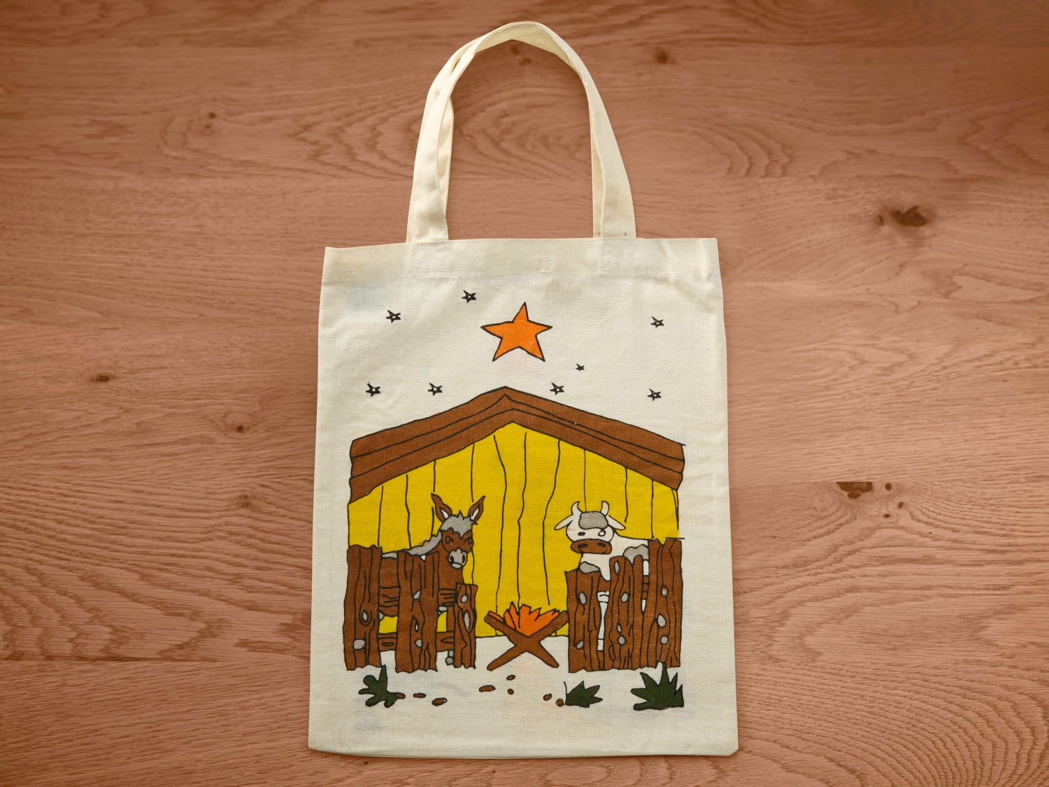 Beige Stofftasche mit aufgedruckter Krippenszene, bestehend aus gelber Stallhütte mit braunem Dach und Zaun, einem Esel links, einer Kuh rechts, brennendem Feuer in der Mitte, orangefarbenem Stern oben und kleinen schwarzen Sternen drumherum.