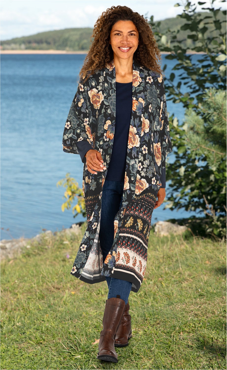 Frau am See trägt lange Viskose-Bluse mit floralem Muster in dunklen Farben, dunkelblaue Jeans, braune Stiefel, lockige Haare, sommerliche Naturkulisse mit Wasser und grüner Wiese im Hintergrund