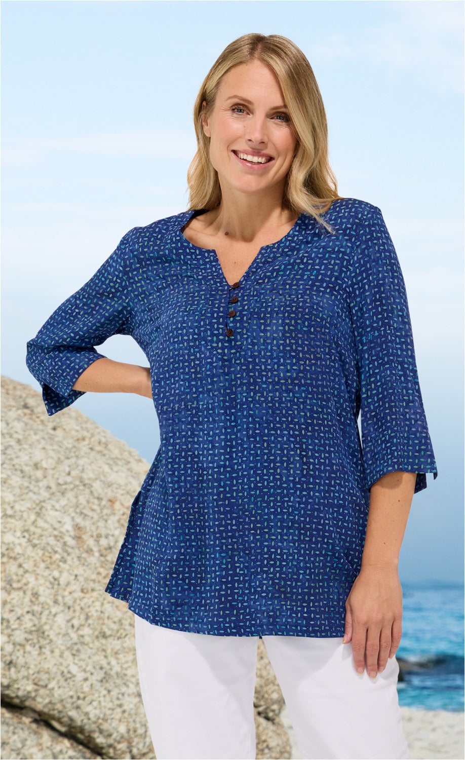 Frau trägt blaue Viskose-Tunika mit kleinem Muster und 3/4-Ärmeln, lächelt, steht vor Felsen am Meer, weiße Hose sichtbar, blauer Himmel im Hintergrund