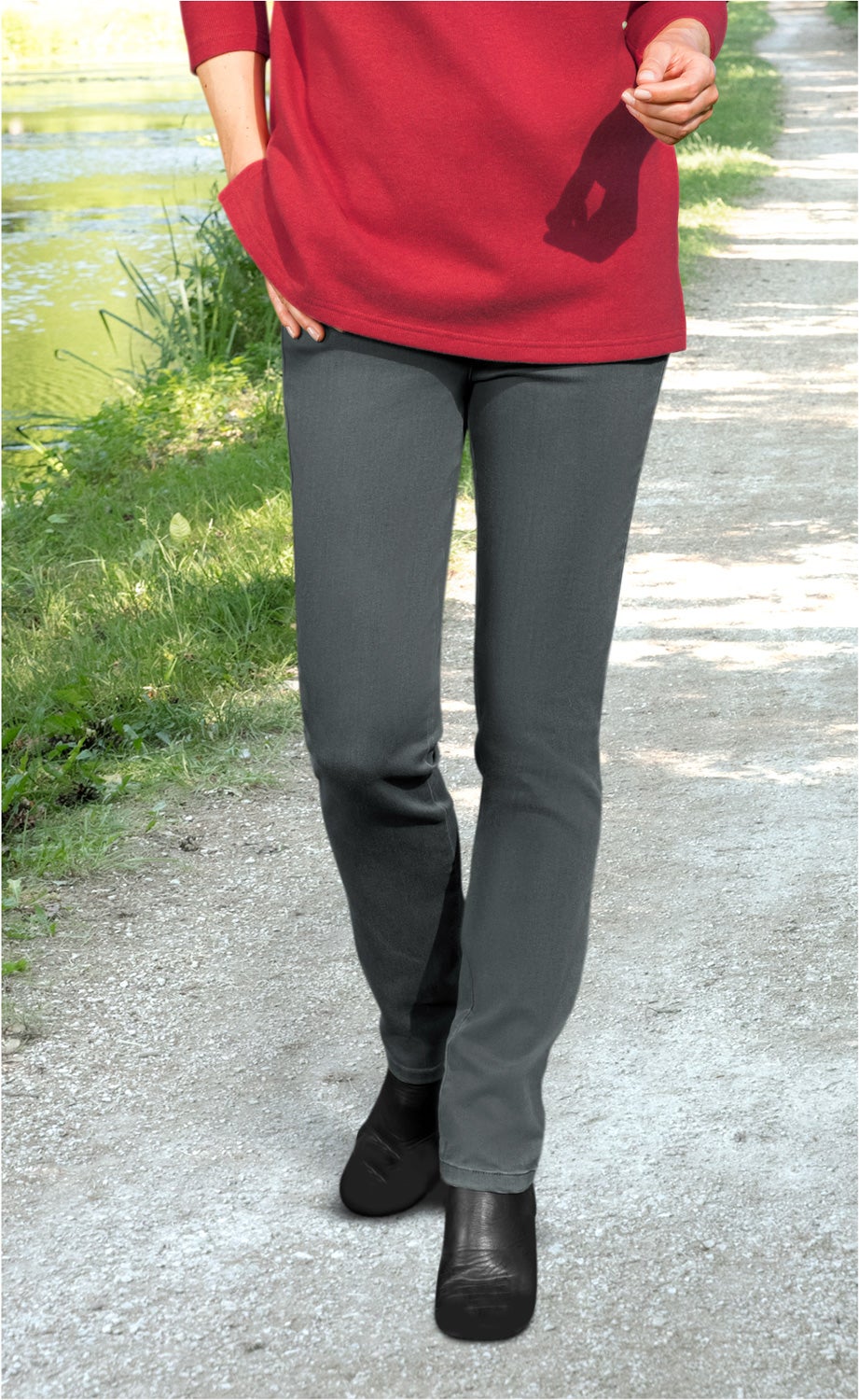 Person geht auf einem Weg neben Wasser, trägt graue Baumwoll-Mix-Hose, schwarzes Schuhwerk und roten Pullover, rechte Hand in Hosentasche, linke Hand leicht erhoben, sonniger Tag, grüne Pflanzen am Wegesrand