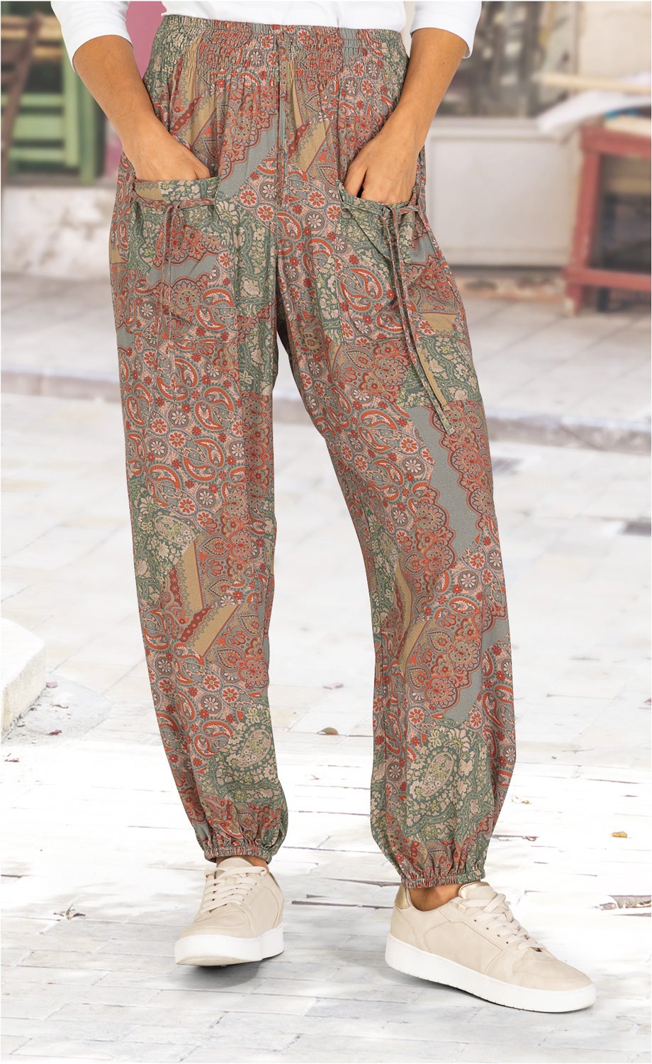 Beine mit Viskose-Hose in buntem Paisley-Muster, elastischem Bund und Bündchen, Hände in Taschen, helle Sneaker, heller Boden, verschwommener Hintergrund eines Außenbereichs