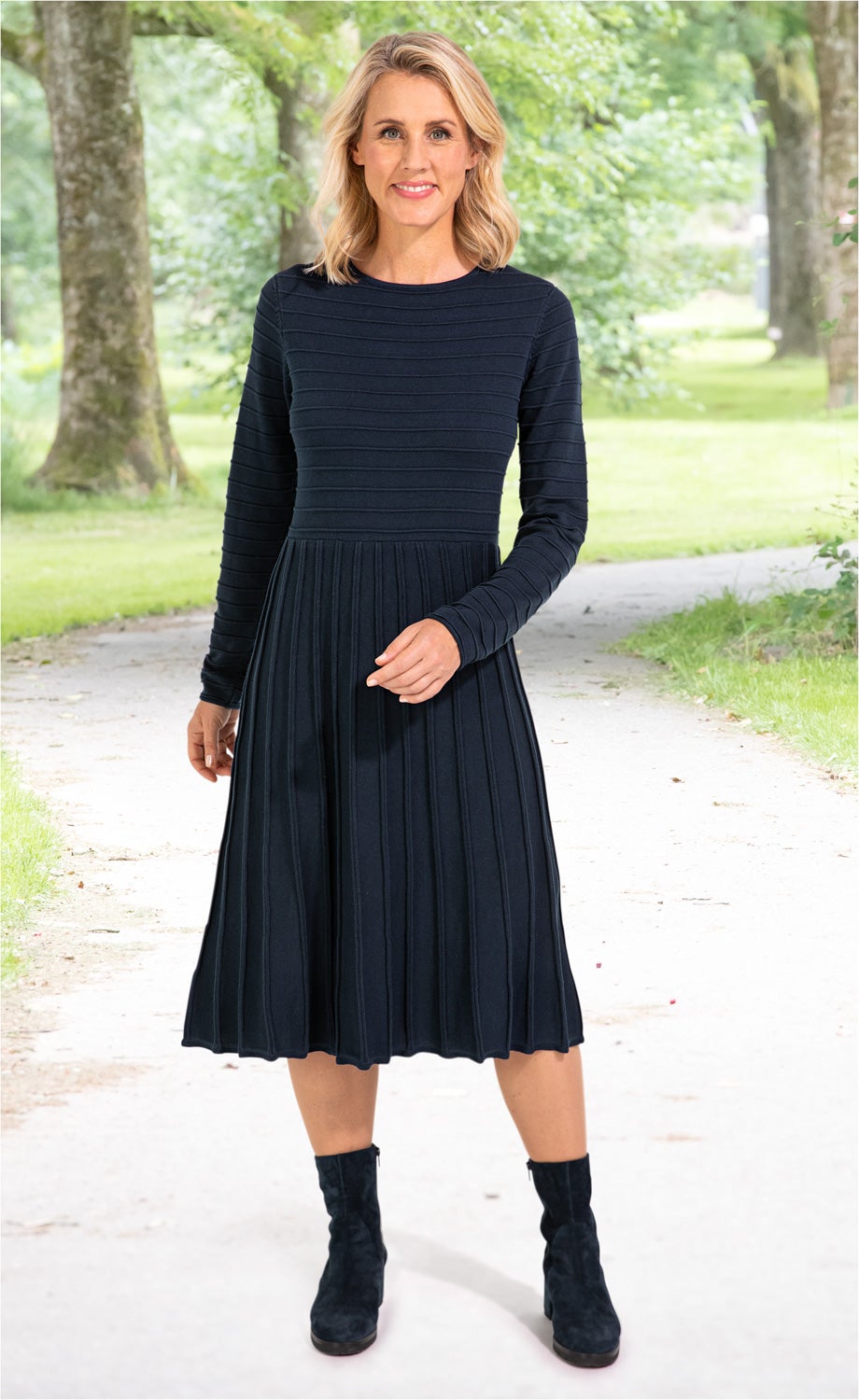Frau trägt navyblaues, langärmliges Baumwoll-Kleid mit feinen Quer- und Längsrippen, knieumspielendem Rock, stehend auf Gehweg mit grünen Bäumen im Hintergrund, kombiniert mit schwarzen Stiefeletten