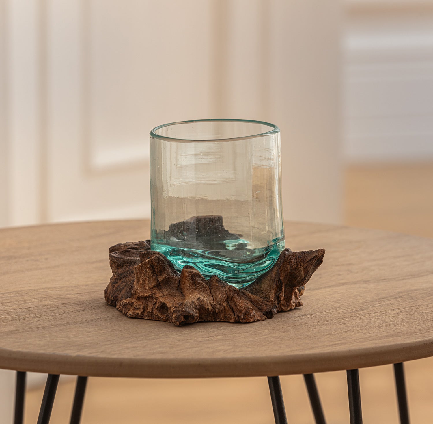 transparentes Windlicht aus Glas mit unregelmäßigem Rand, sitzt auf einem braunen, naturbelassenen Wurzelholzring, steht auf rundem Holztisch mit schwarzen Metallbeinen, heller Hintergrund mit Türen und Wänden unscharf sichtbar
