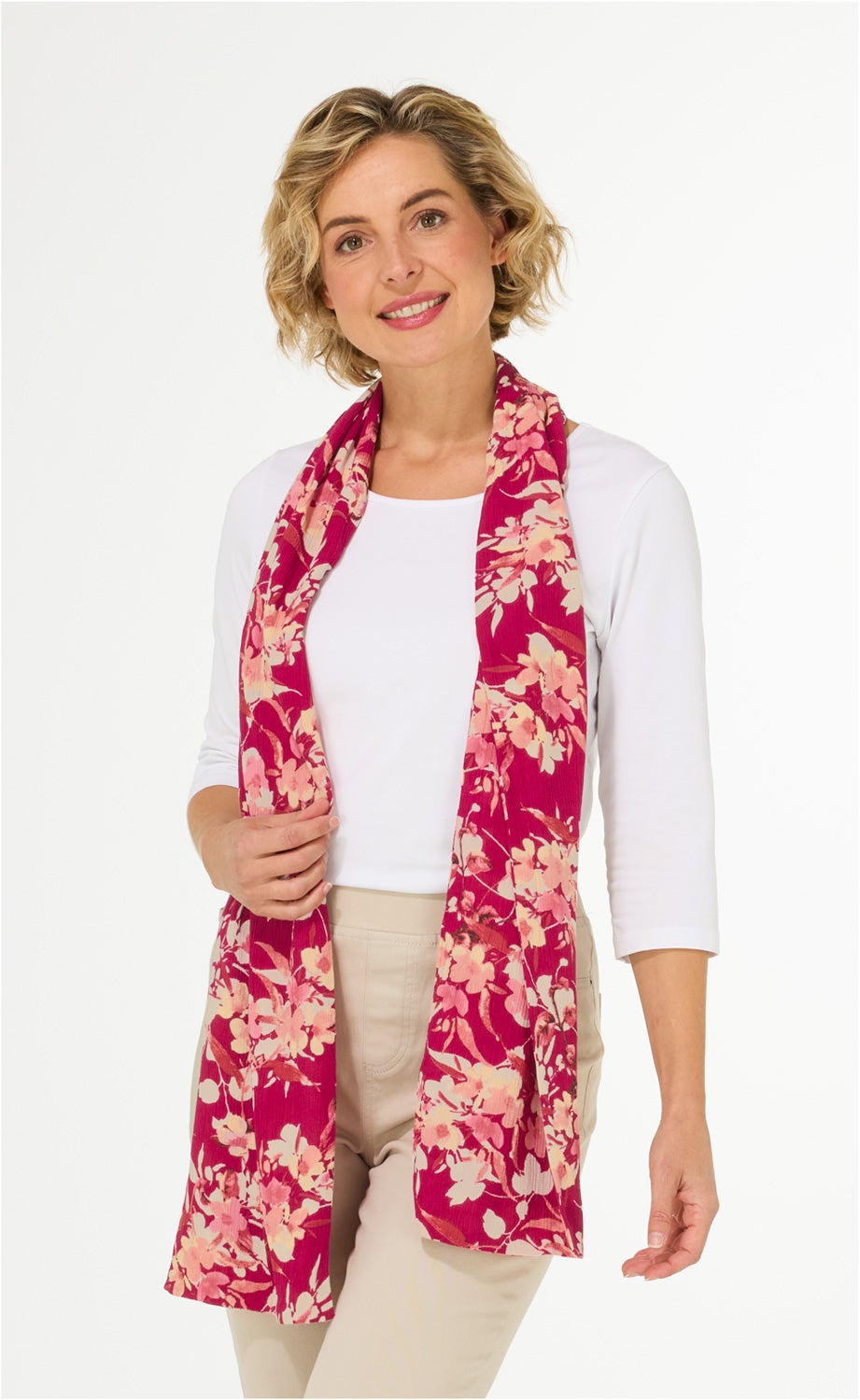 Frau mit kurzem blonden Haar trägt weißen 3/4-Arm-Shirt, beige Hose und pink-roten Viskose-Schal mit floralem Muster, hält Schal locker vor dem Körper, lächelt freundlich, vor hellem Hintergrund.