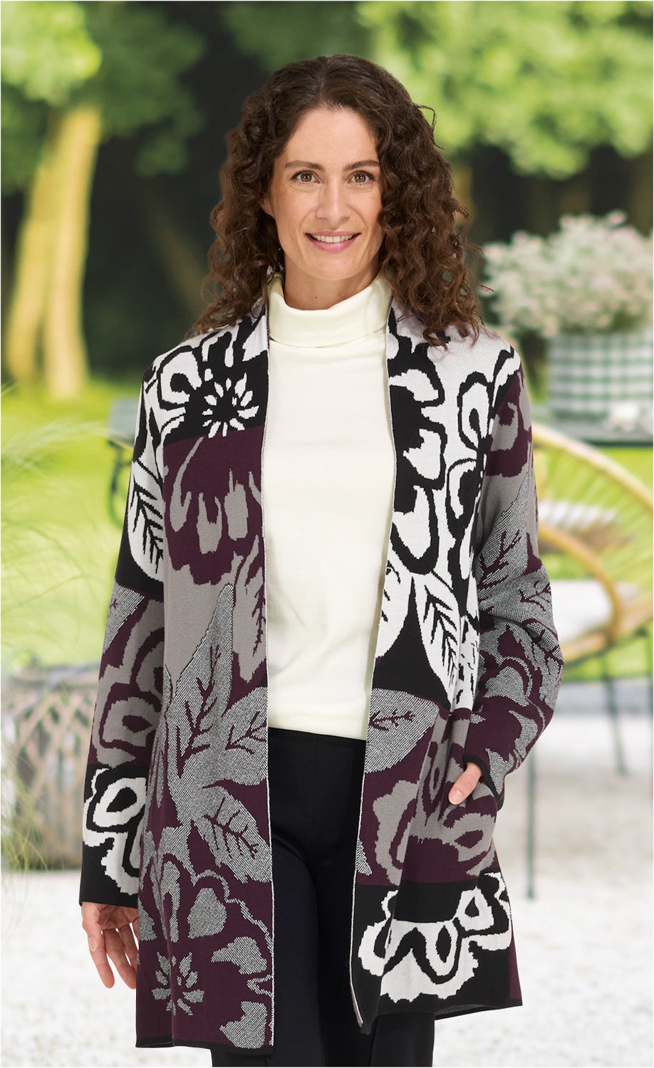 Frau trägt Baumwoll-Mix-Strickjacke mit großem floralen Muster in Schwarz, Grau und Dunkelrot, kombiniert mit weißem Rollkragenpullover und schwarzer Hose, steht vor unscharf grünem Gartenhintergrund