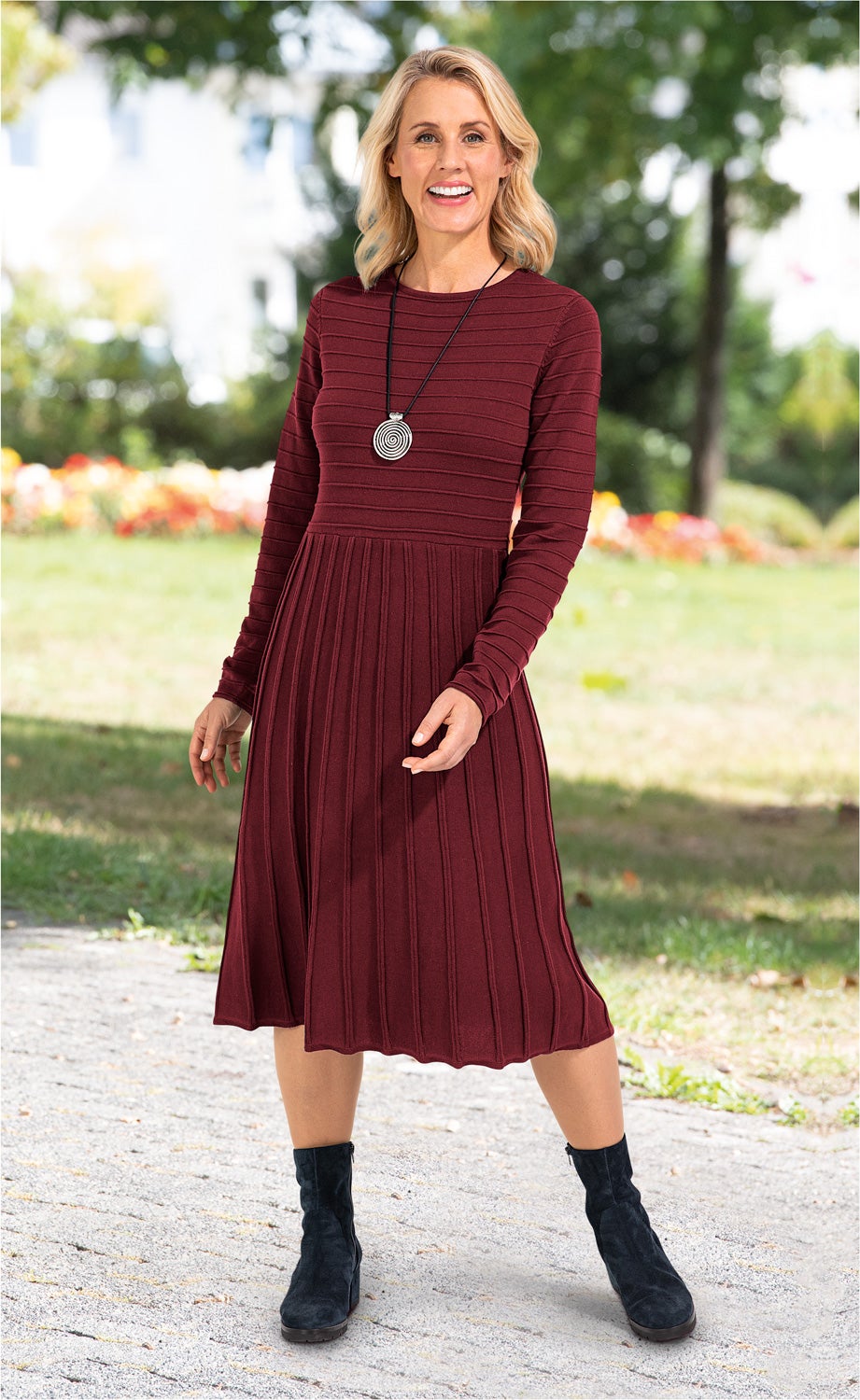 Frau trägt bordeauxrotes, langärmeliges Baumwoll-Kleid mit vertikalen Streifen, knielang, dazu schwarze Stiefeletten und silberne Spiral-Halskette, steht auf gepflastertem Weg, grüner Parkhintergrund, lächelt