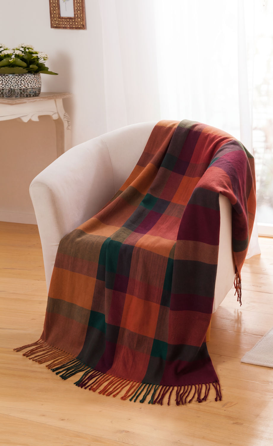 Alpaka-Decke mit kariertem Muster in warmen Herbstfarben, rot, orange, grün und violett, mit Fransen, auf einem weißen Sessel in hellem Raum mit Holzboden drapiert