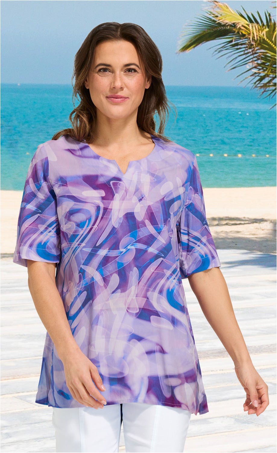 Frau trägt helle Viskose-Tunika mit abstraktem blau-lila Muster, halblange Ärmel, Rundhalsausschnitt mit kleinem Schlitz, weiße Hose, Strand mit Meer und Palme im Hintergrund