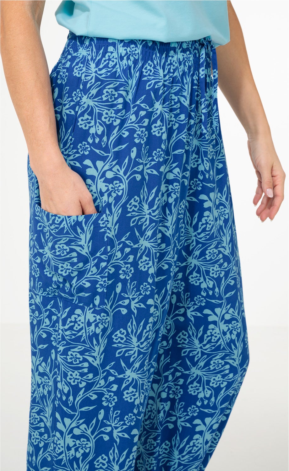Blaue Viskose-Hose mit hellblauem floralem Muster, elastischem Bund und seitlicher Tasche, getragen von Person mit Hand in der Tasche und hellem Oberteil