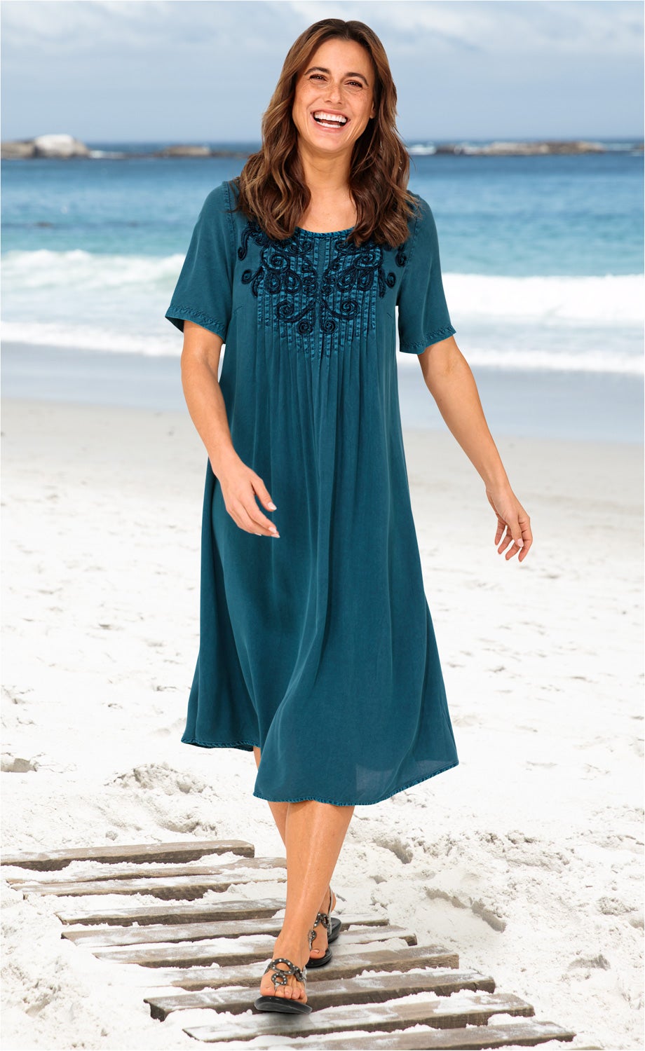 Frau läuft am Strand auf Holzsteg, trägt blaues Viskose-Kleid mit kurzem Arm und floraler Stickerei am Ausschnitt, schwarze Sandalen, lächelt, Hintergrund Meer mit Wellen und bewölktem Himmel