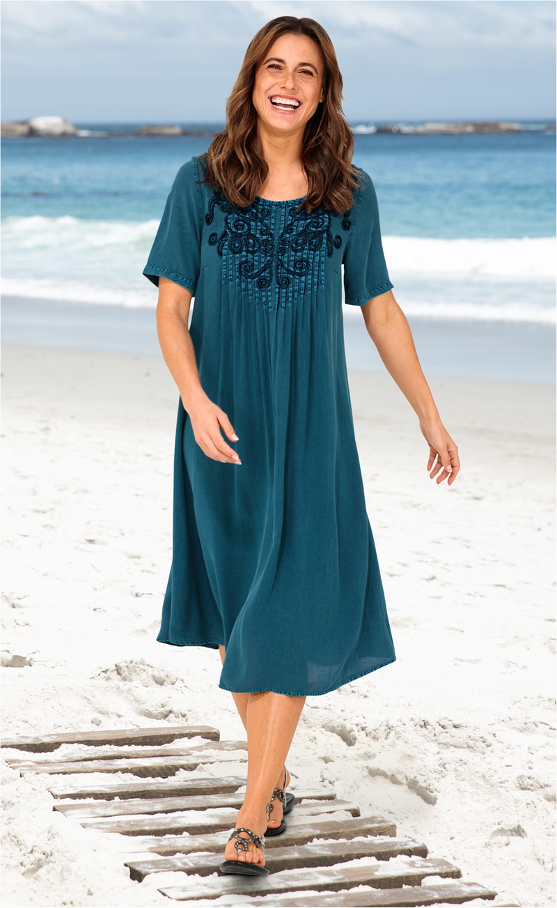 Frau läuft am Strand auf Holzsteg, trägt blaues Viskose-Kleid mit kurzem Arm und floraler Stickerei am Ausschnitt, schwarze Sandalen, lächelt, Hintergrund Meer mit Wellen und bewölktem Himmel