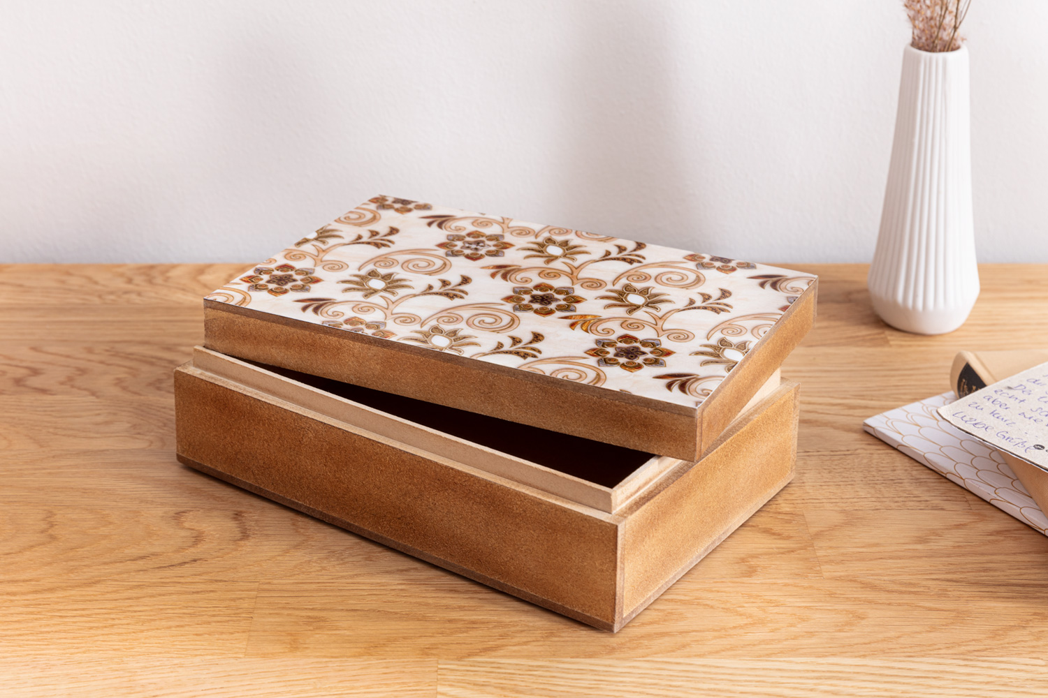 Halboffene rechteckige Holzbox mit hellbraunen Seiten und einem Deckel, der mit einem weißen, floral und ornamentartig verzierten Muster dekoriert ist, steht auf einem hellen Holztisch neben einem weißen, hohen, geriffelten Keramikvase.