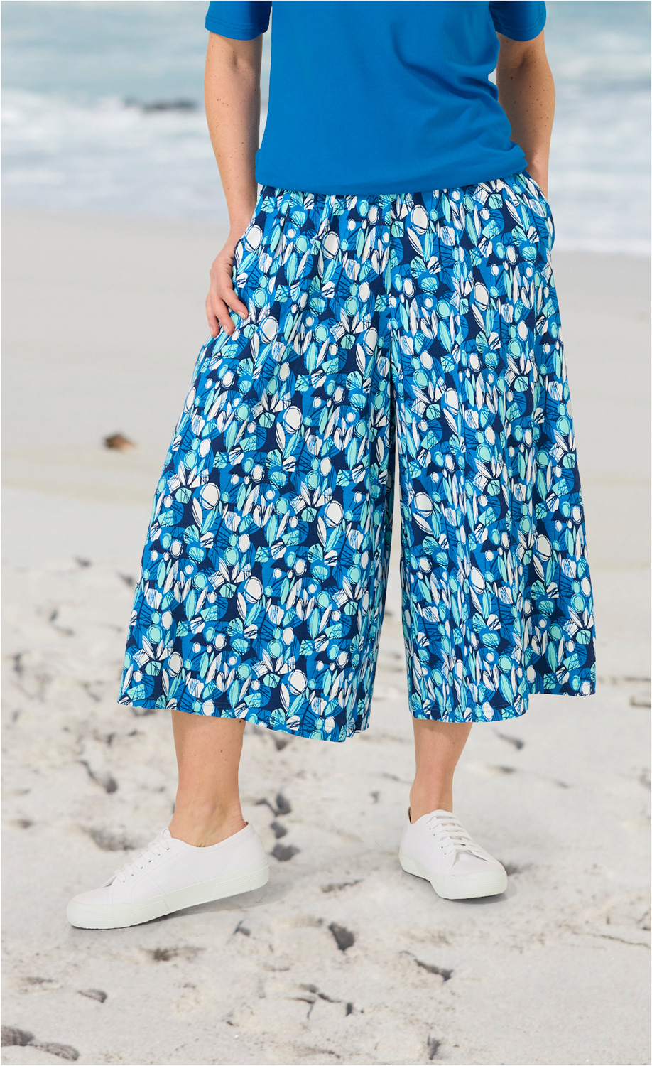 Viskose-Mix-Hosenrock mit blau-weißem floralem Muster, weite Passform, knielang, elastischer Bund, kombiniert mit blauem Oberteil und weißen Sneakern, stehend am Strand mit sandigem Hintergrund