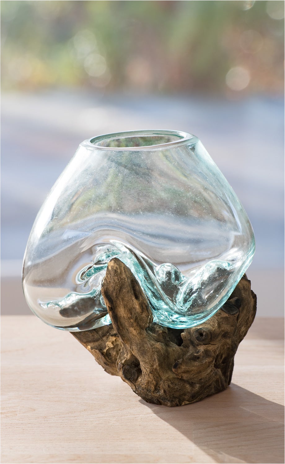 Transparente Glasvase mit geschwungener Form, die auf einem naturbelassenen, knorrigen Holzsockel sitzt, auf hellem Untergrund vor unscharfem, mehrfarbigem Hintergrund.