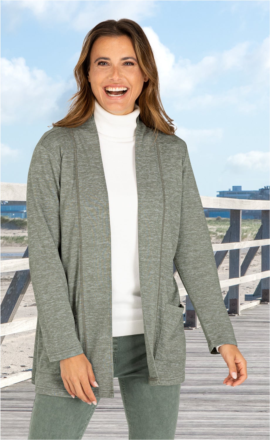 Frau trägt grüne Viskose-Mix-Jacke mit aufgesetzten Taschen über weißem Rollkragenpullover, steht auf Holzsteg am Strand, blauer Himmel mit Wolken im Hintergrund, lächelt freundlich