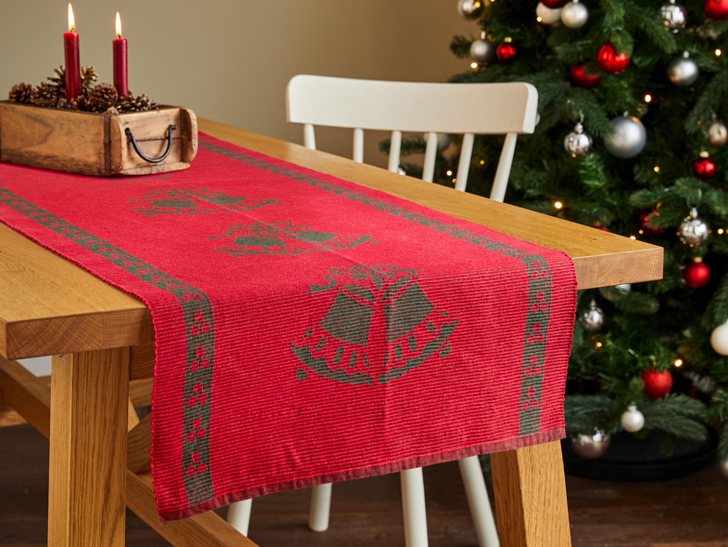 Roter Baumwoll-Tischläufer mit grünen Glockenmotiven auf hellem Holztisch, dekorative Holzbox mit roten Kerzen und Tannenzapfen, weißer Stuhl und geschmückter Weihnachtsbaum im Hintergrund