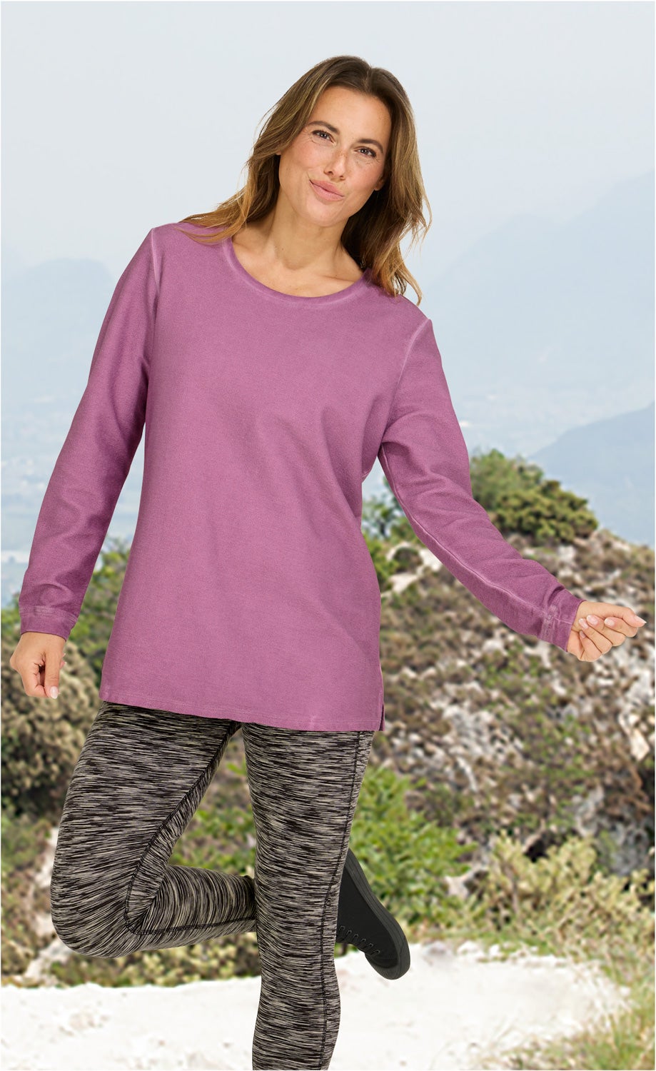 Frau trägt langärmliges, violettes Baumwoll-Shirt, kombiniert mit grauen, gemusterten Leggings, steht auf einem Bergpfad vor Naturkulisse, hebt ein Bein leicht an, leicht lächelnd, entspannte Pose