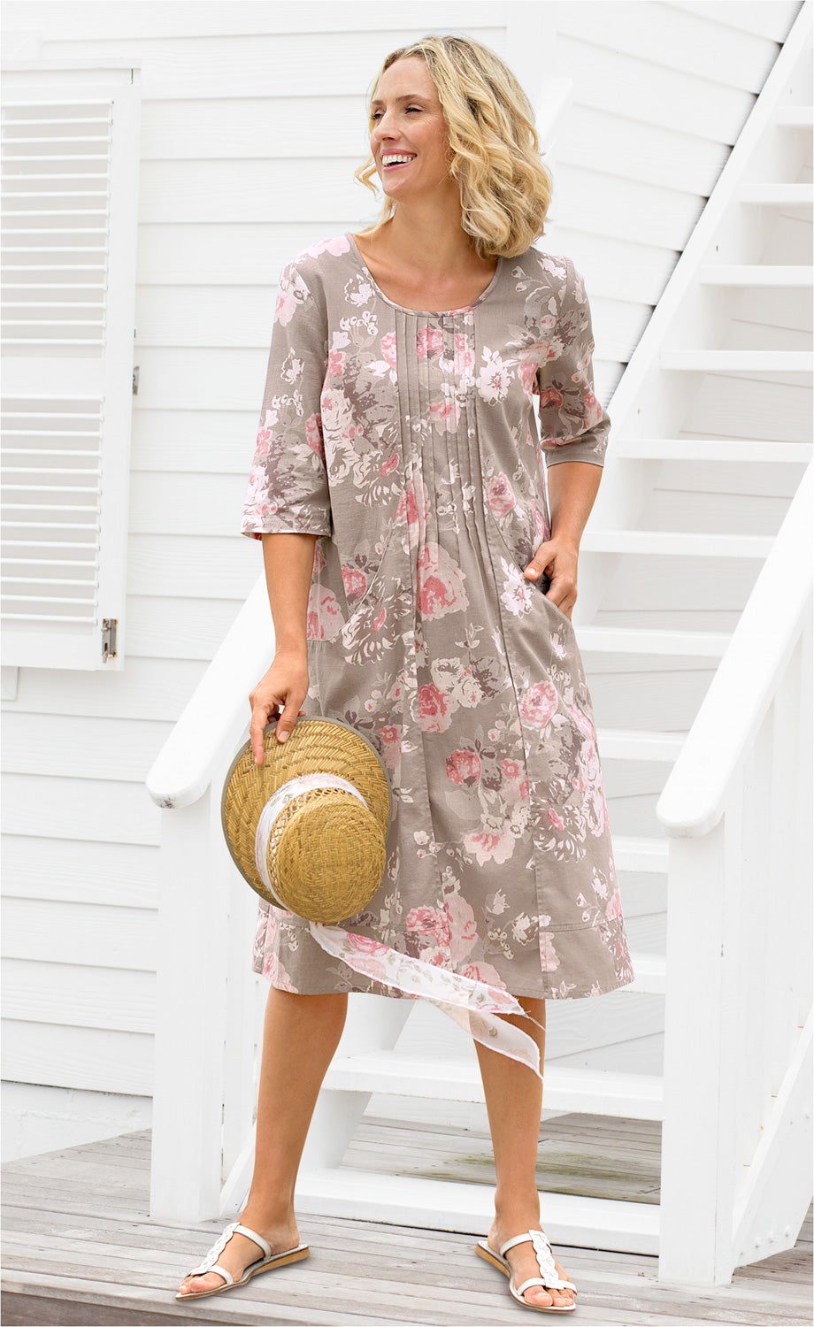 Frau trägt beige Baumwoll-Mix-Kleid mit floralem Muster in rosa und weiß, 3/4-Ärmeln, hält Strohhut mit weißem Band, weiße Sandalen, steht vor weißem Haus mit Holztreppe und Holzveranda
