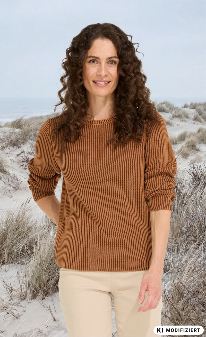 Frau mit langen lockigen braunen Haaren trägt braunen gestreiften Pullover und helle Hose, steht am Strand mit Sanddünen und grauem Himmel im Hintergrund, lächelt freundlich