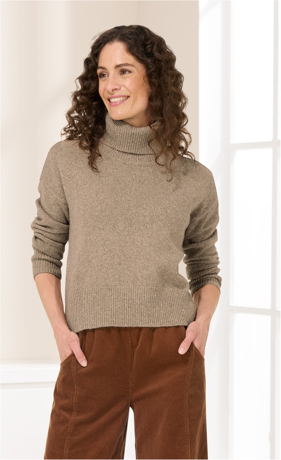 Frau mit lockigen dunklen Haaren trägt beige Woll-Mix-Pullover mit Rollkragen und braune Cordhose, steht vor hellem Fenster, Hände in Taschen, lächelt und schaut nach rechts