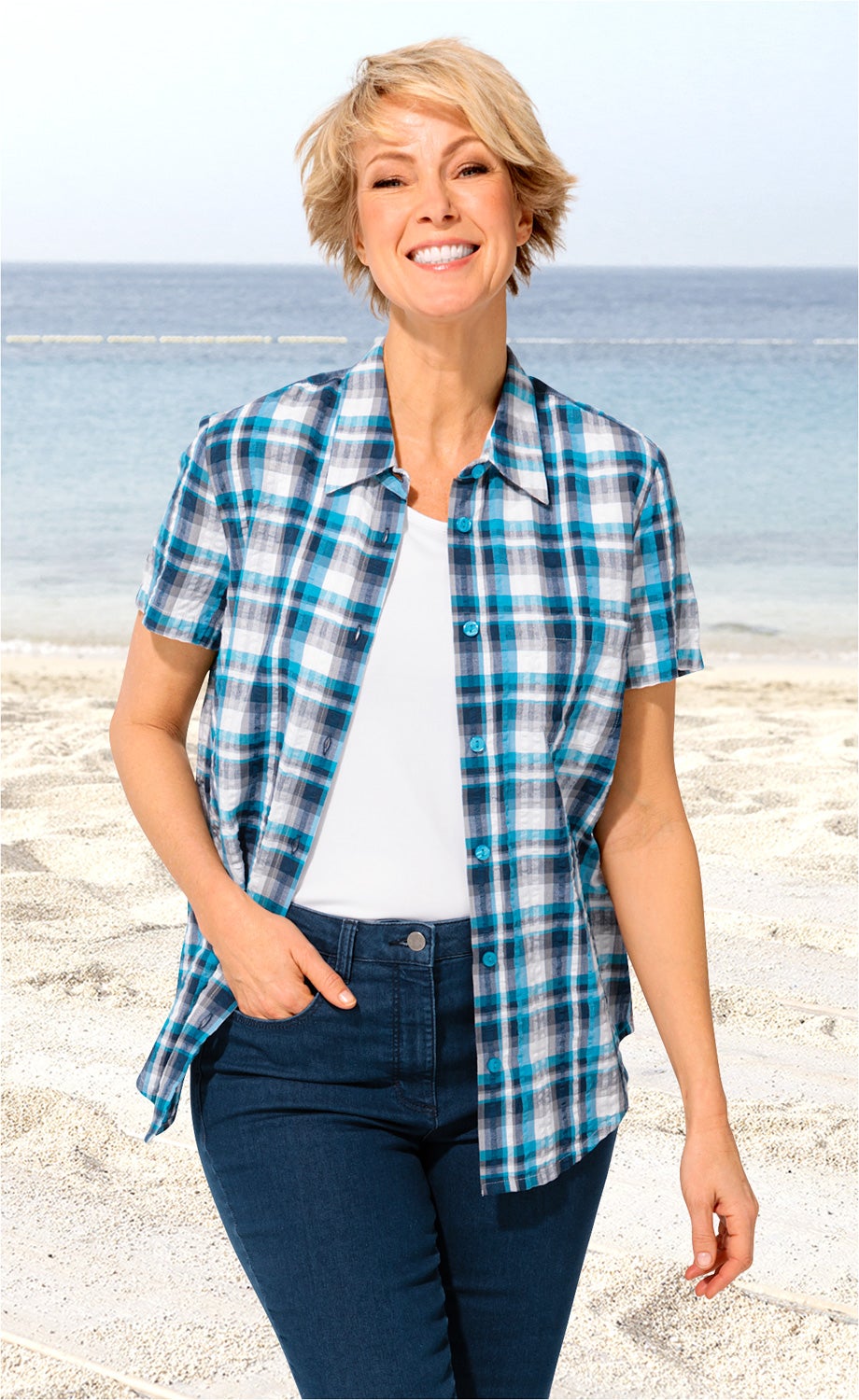 Frau am Strand trägt kurzärmlige, karierte Baumwoll-Bluse in Blau- und Grautönen, weiße Unterzieh-Top und dunkle Jeanshose, lächelt freundlich, Hand in Tasche, Meer im Hintergrund.