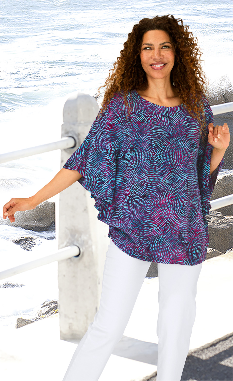 Frau trägt Viskose-Bluse mit blau-lila Punktmuster, steht am Geländer am Meer, lockige Haare, weiße Hose, lächelnd, entspannte Pose, helle, sonnige Umgebung