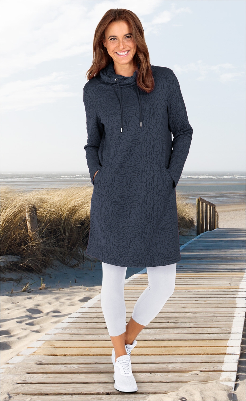 Frau trägt dunkelblaues Viskose-Mix-Kleid mit Stehkragen und Kordel, weiße Leggings und weiße Sneaker, steht lächelnd auf Holzsteg am Strand, im Hintergrund Dünen mit Strandgras und Meer unter leicht bewölktem Himmel