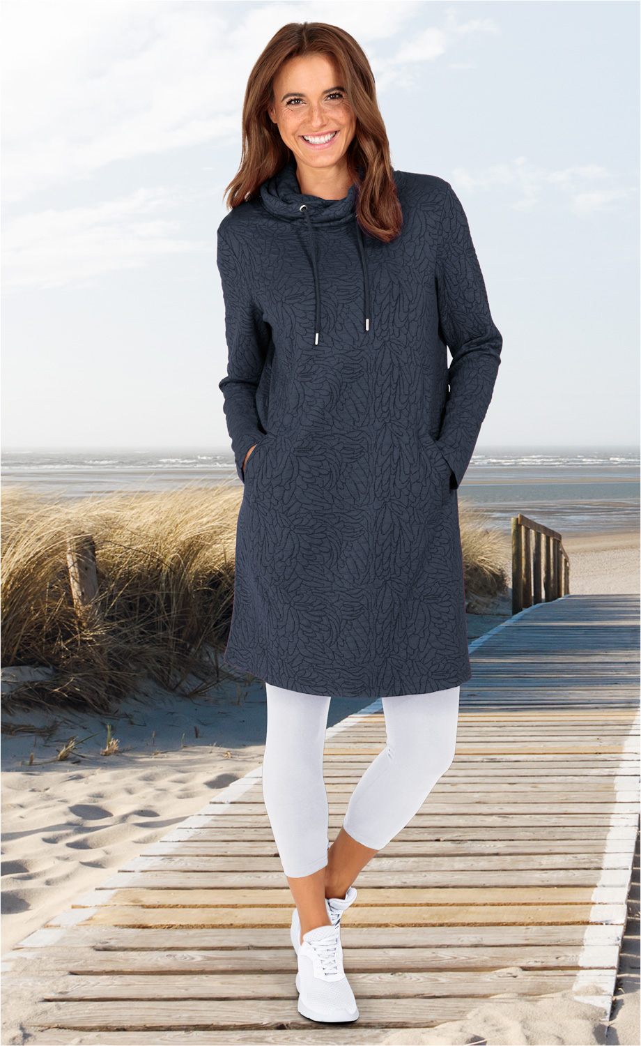Frau trägt dunkelblaues Viskose-Mix-Kleid mit Stehkragen und Kordel, weiße Leggings und weiße Sneaker, steht lächelnd auf Holzsteg am Strand, im Hintergrund Dünen mit Strandgras und Meer unter leicht bewölktem Himmel