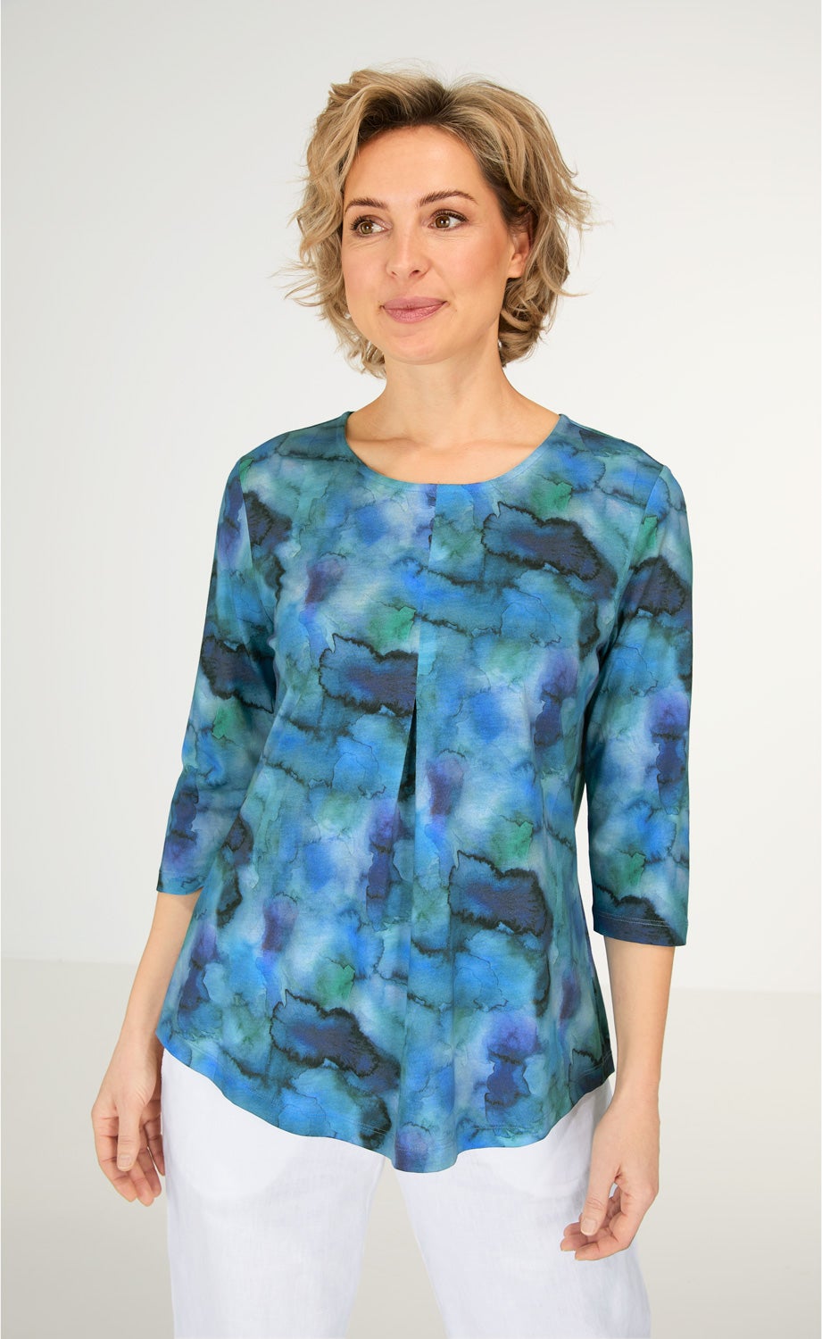 Frau trägt blau-grün gemustertes Baumwoll-Mix Shirt mit 3/4-Ärmeln und Rundhalsausschnitt, stehende Falte vorne, kombiniert mit weißer Hose, neutraler Hintergrund