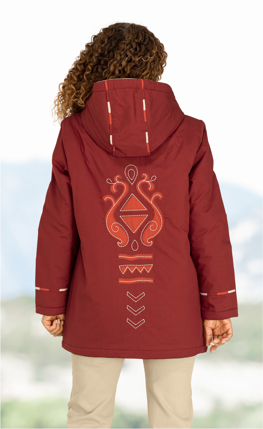 Person von hinten mit langen lockigen Haaren, trägt rote Kapuzenjacke mit hellrotem, geometrisch-floralem Sticker auf dem Rücken, helle Hose, helle und unscharfe Hintergrundlandschaft, Arme hängen locker herunter