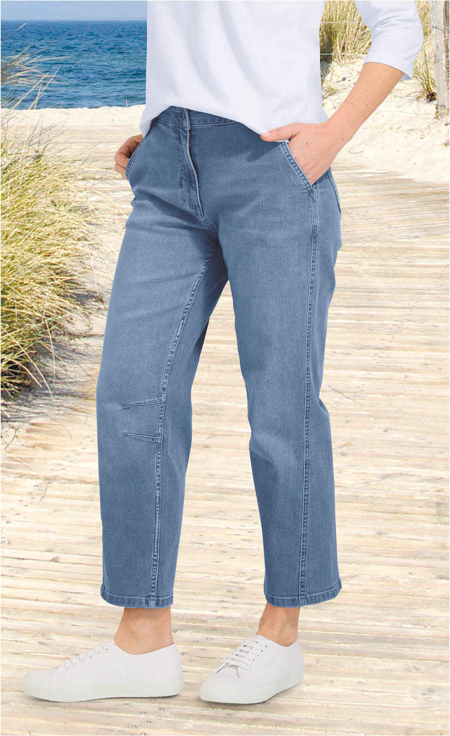Frau trägt blaue Baumwoll-Mix-Hose mit geradem Bein und seitlichen Eingrifftaschen, kombiniert mit weißen Sneakern, stehend auf hellem Holzsteg am Strand mit Meer und Dünengras im Hintergrund.