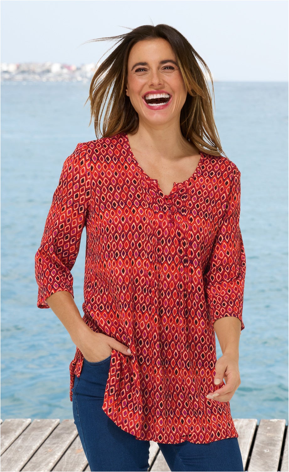 Frau am Steg trägt rote Viskose-Bluse mit geometrischem Muster, 3/4-Ärmeln und V-Ausschnitt, lächelt, im Hintergrund Meer und Himmel, dunkle lange Haare wehen im Wind, Hand in Hosentasche, Blaue Hose