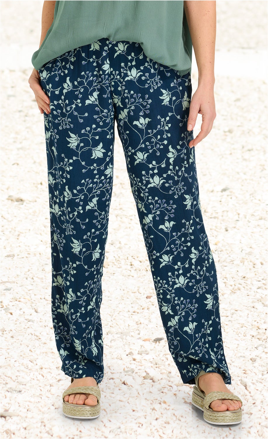 Damen Viskose-Hose dunkelblau mit hellgrünem floralem Muster, gerade geschnitten, kombiniert mit grüner Bluse und beige-geflochtenen Sandalen, stehend auf heller Kiesfläche