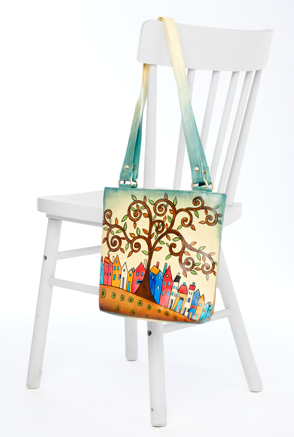 Handbemalte Leder-Handtasche mit farbenfrohem Motiv eines geschwungenen Baums und bunter Häuserreihe, hängt an der Lehne eines weißen Holzstuhls, Träger verlaufend in Grün- und Gelbtönen
