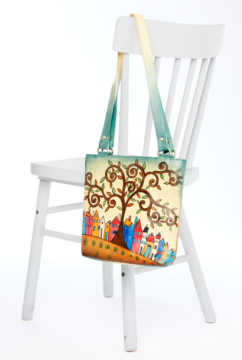 Handbemalte Leder-Handtasche mit farbenfrohem Motiv eines geschwungenen Baums und bunter Häuserreihe, hängt an der Lehne eines weißen Holzstuhls, Träger verlaufend in Grün- und Gelbtönen