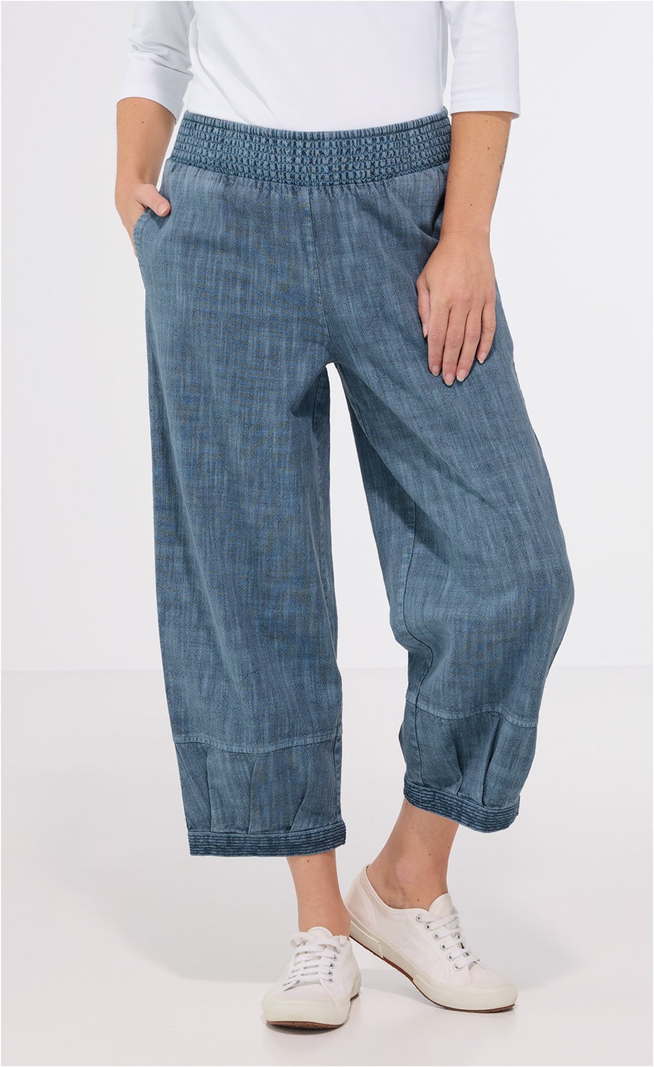 Baumwoll-Hose in blau mit weitem Bein und gesmoktem Bund, Saum mit Ziernähten, getragen von Person mit weißem Oberteil und weißen Sneakern, rechte Hand in Hosentasche, linker Arm locker am Bein.