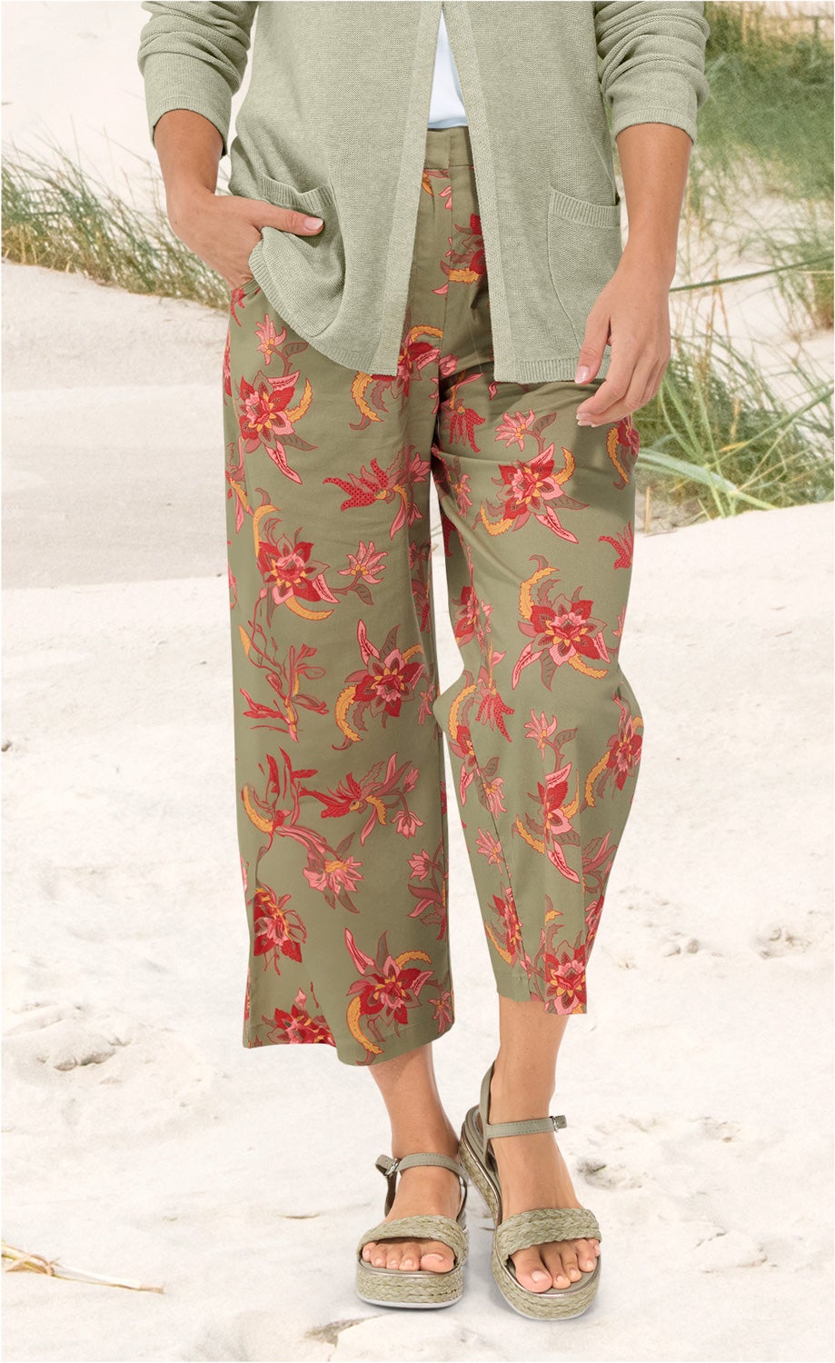 Baumwoll-Mix-Hose in khaki mit weitem Bein und floralem Muster in rot, pink und gelb, kombiniert mit grünem Strickcardigan, Pop-up-Sandalen mit geflochtenem Riemen, Strand und Dünen im Hintergrund